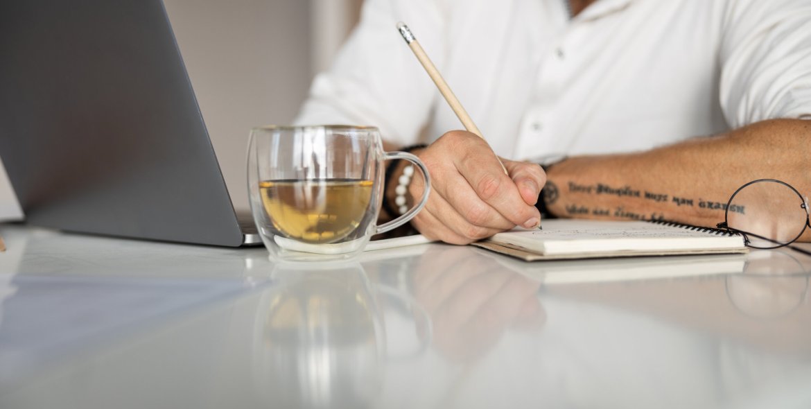Cropped photo of a white-collar employee putting his thoughts on paper with a laptop and tea by his side